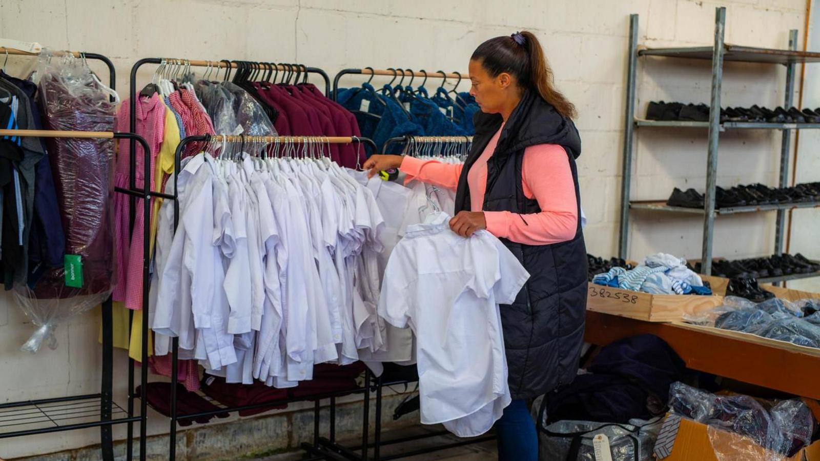 Pia Honey is looking through a rack of children's school shirts in a warehouse. Other clothing can be seen behind and there are also racks of shoes