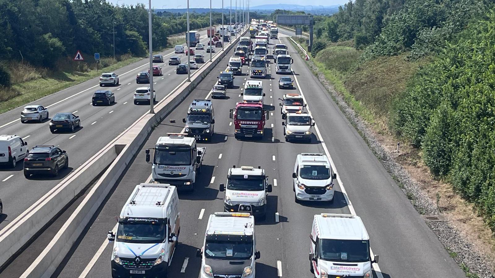 Hundreds of lorries and vans are in convoy along three lanes of a motorway, stretching back as far as the eye can see. They have their lights on