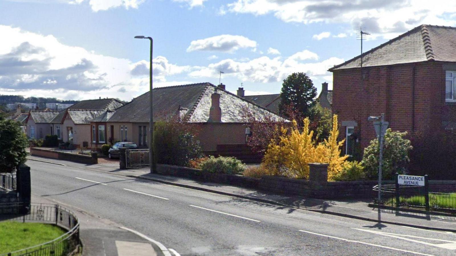 A town street with a sign in the foreground and two brown brick houses with trees cars in the driveways and a blue cloudy sky on a sunny day