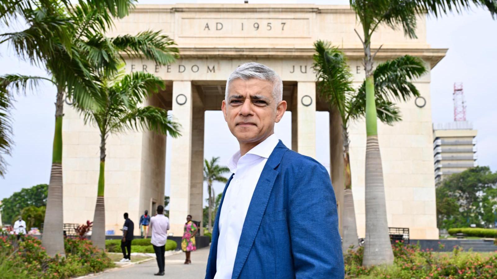 Sadiq Khan stands in front of a ceremonial arch. He is wearing a blue suit and white shirt. There are two rows of palm trees in front of the arch. 