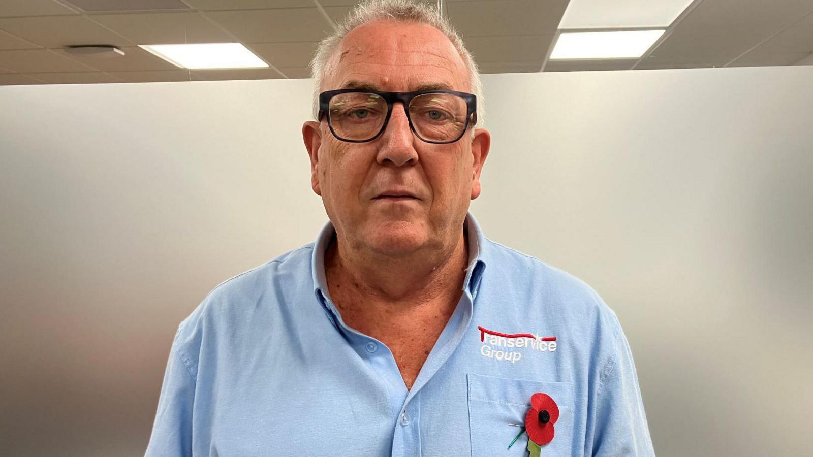 A man stands inside an office building. The man has grey hair and he is wearing glasses. He is wearing a light blue shirt and a red rememberance poppy badge.