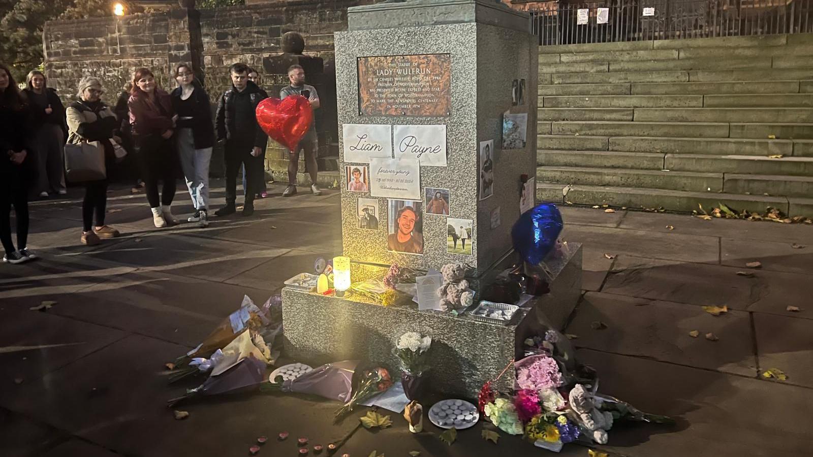 About nine people. mostly women, stand at the foot of a statue.  The base has a paper sign that says "Liam Payne" with pictures and heart shaped balloons placed around the base, candles, flowers and other tributes have been placed on the ground next to it. 