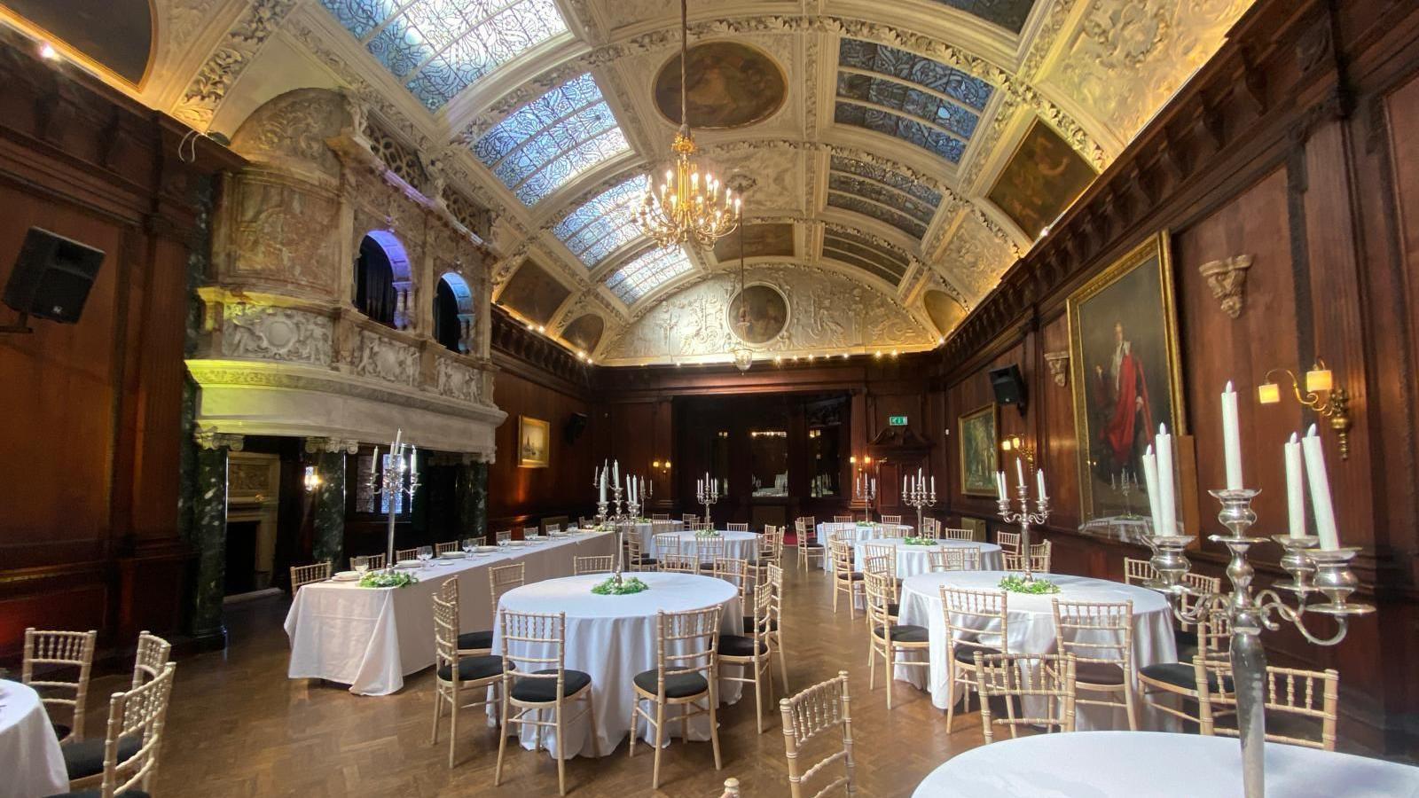 A grand dining room in an old manor. It has ornate decorations on the walls, a big oil painting of a man in a cloak and a number of tables and chairs set up.
