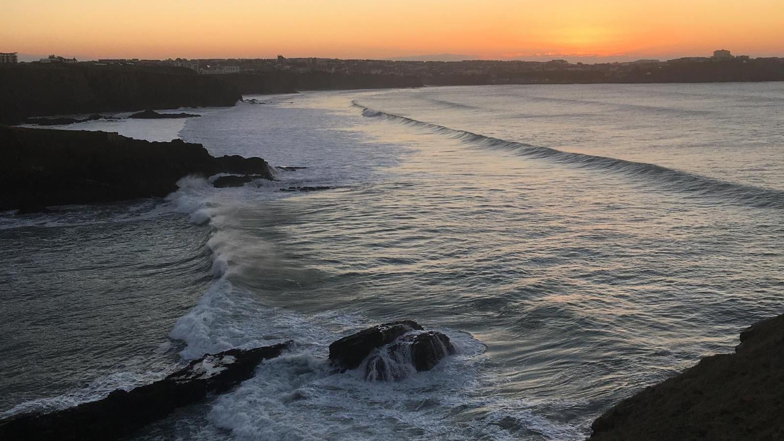 A Cornish beach at sunset. Waves crash in over rocks beneath high cliffs as the yellow sun casts long shadows