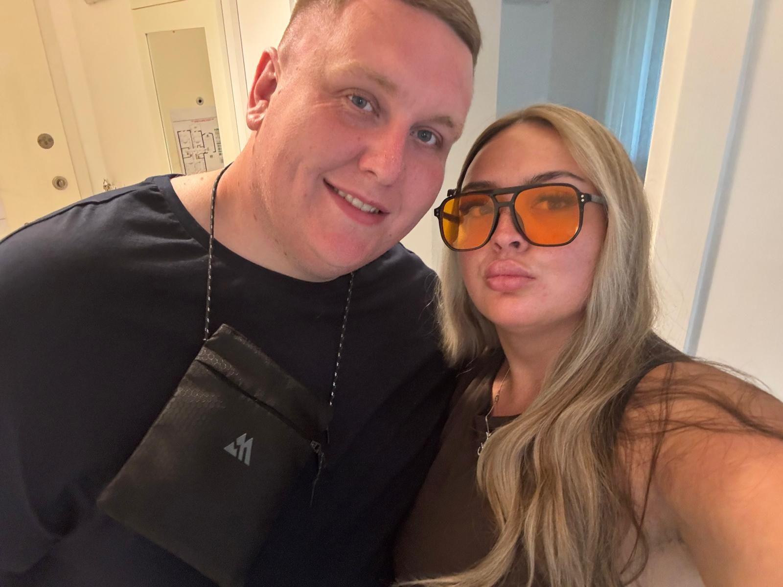 Woman with blonde hair and orange lens sunglasses looks at the camera. A blonde man wearing a black top and a cross body bag is standing next to her