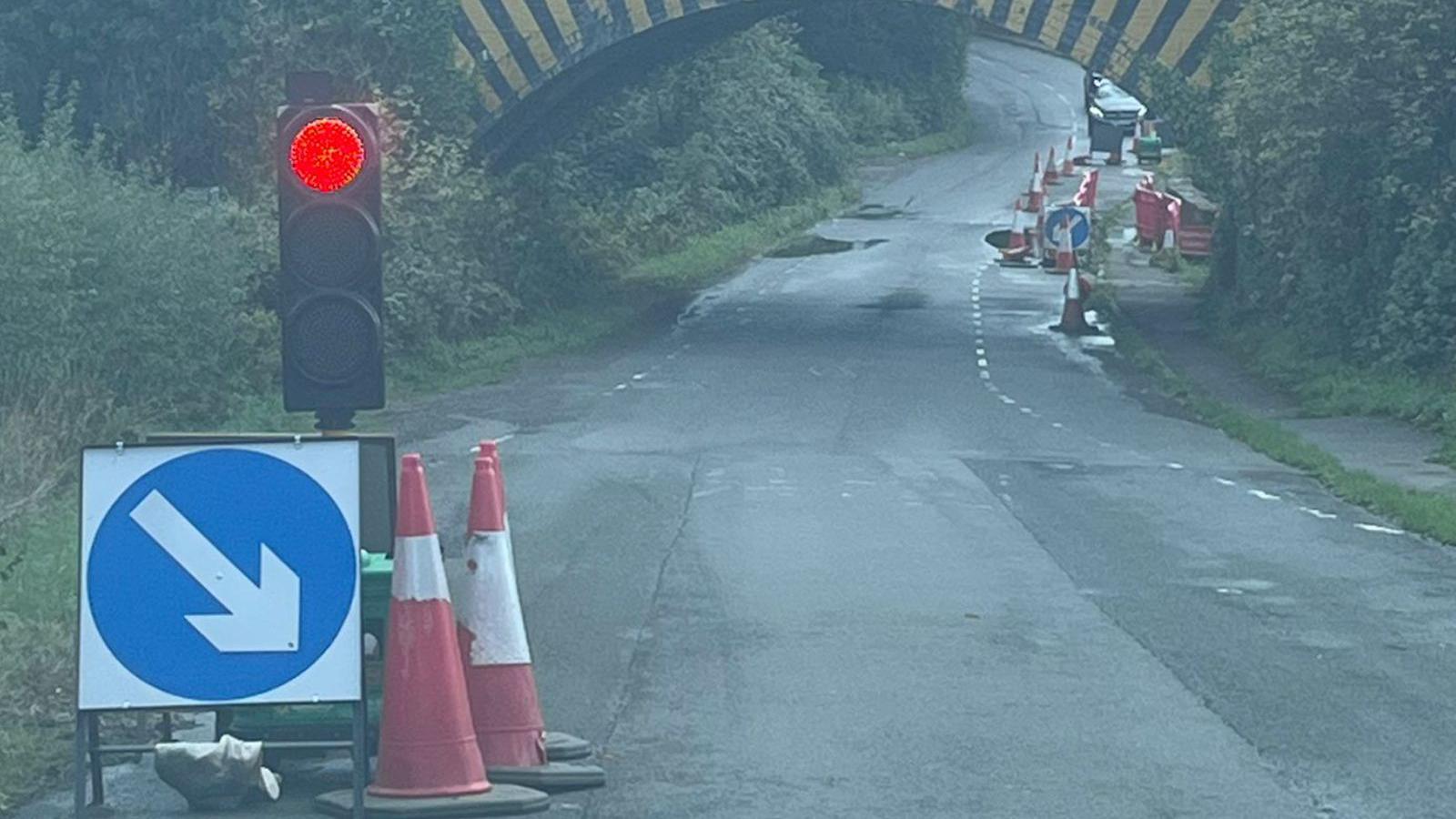 A temporary traffic light holding traffic on a road. In the background is the railway bridge with cars waiting on the other side.