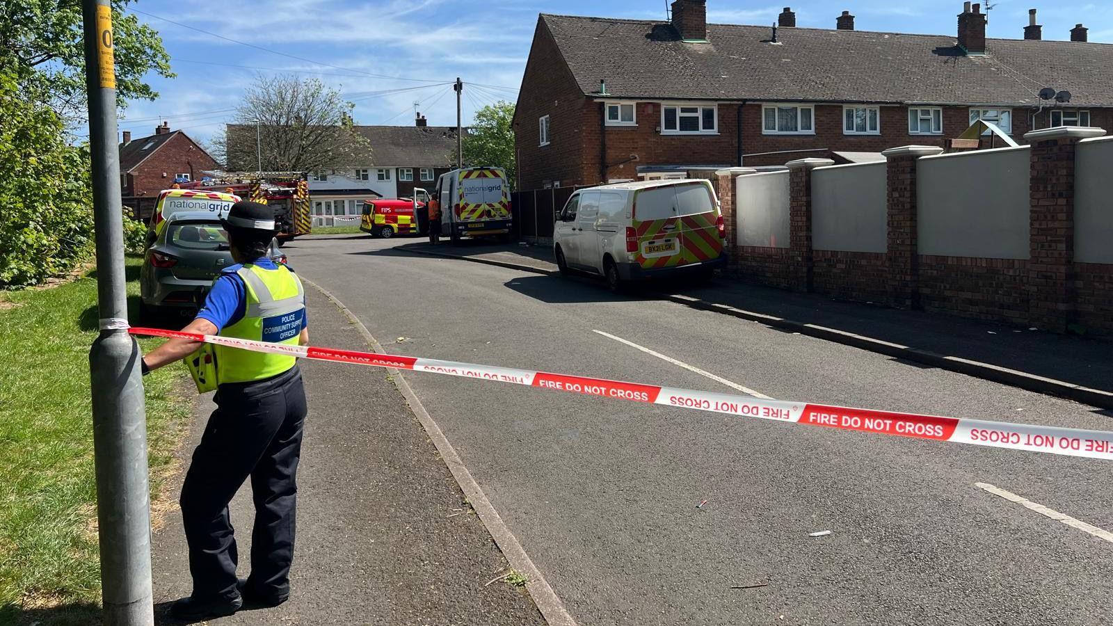 A red and white cordon across a residential street. The cordon says: FIRE DO NOT CROSS. A police officer stands by the cordon.