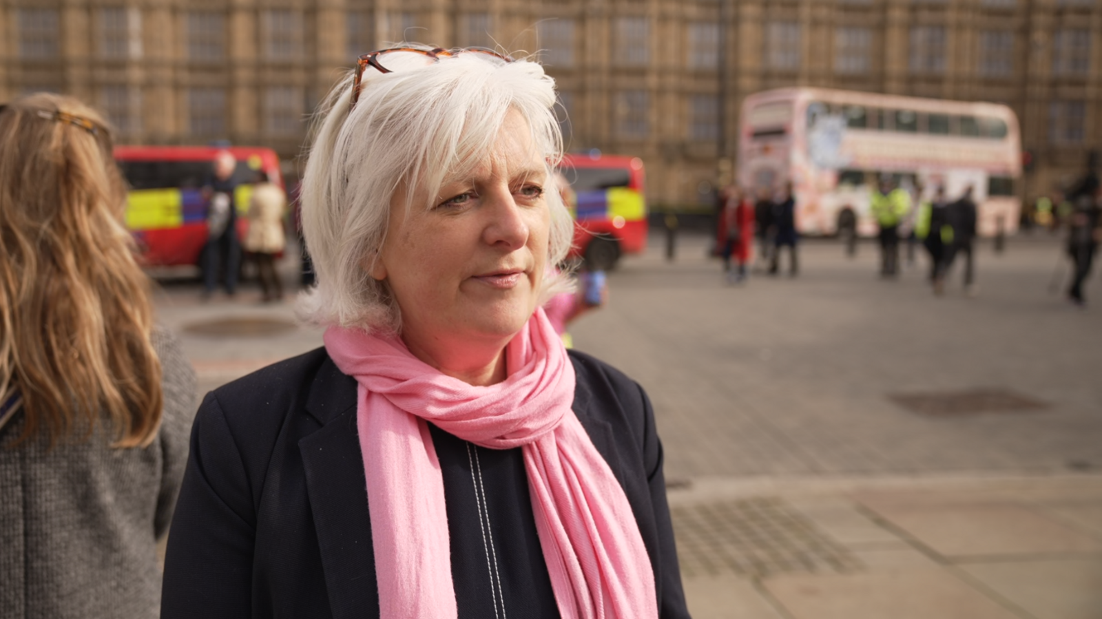 A grey-haired woman with glasses on her head, wearing a pink scarf and black jacket stands in front of parliament.