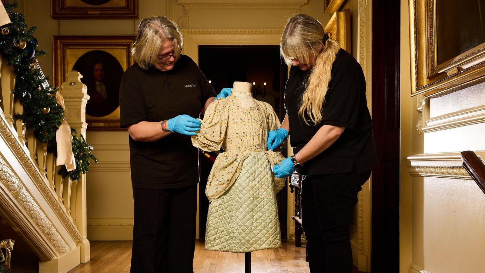 Two women in black uniforms and blue gloves touch a child's dress on a mannequin in the hall of a stately home.