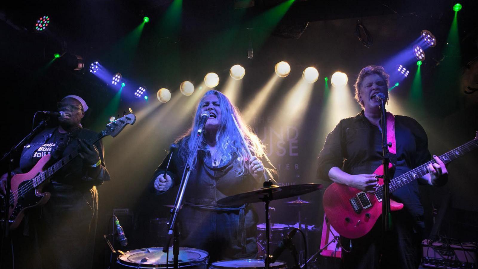 A photograph of the Boilers band playing on stage. Chrissie (left) plays bass, Alison (middle) sings and plays drums, and there is a woman playing guitar on the right