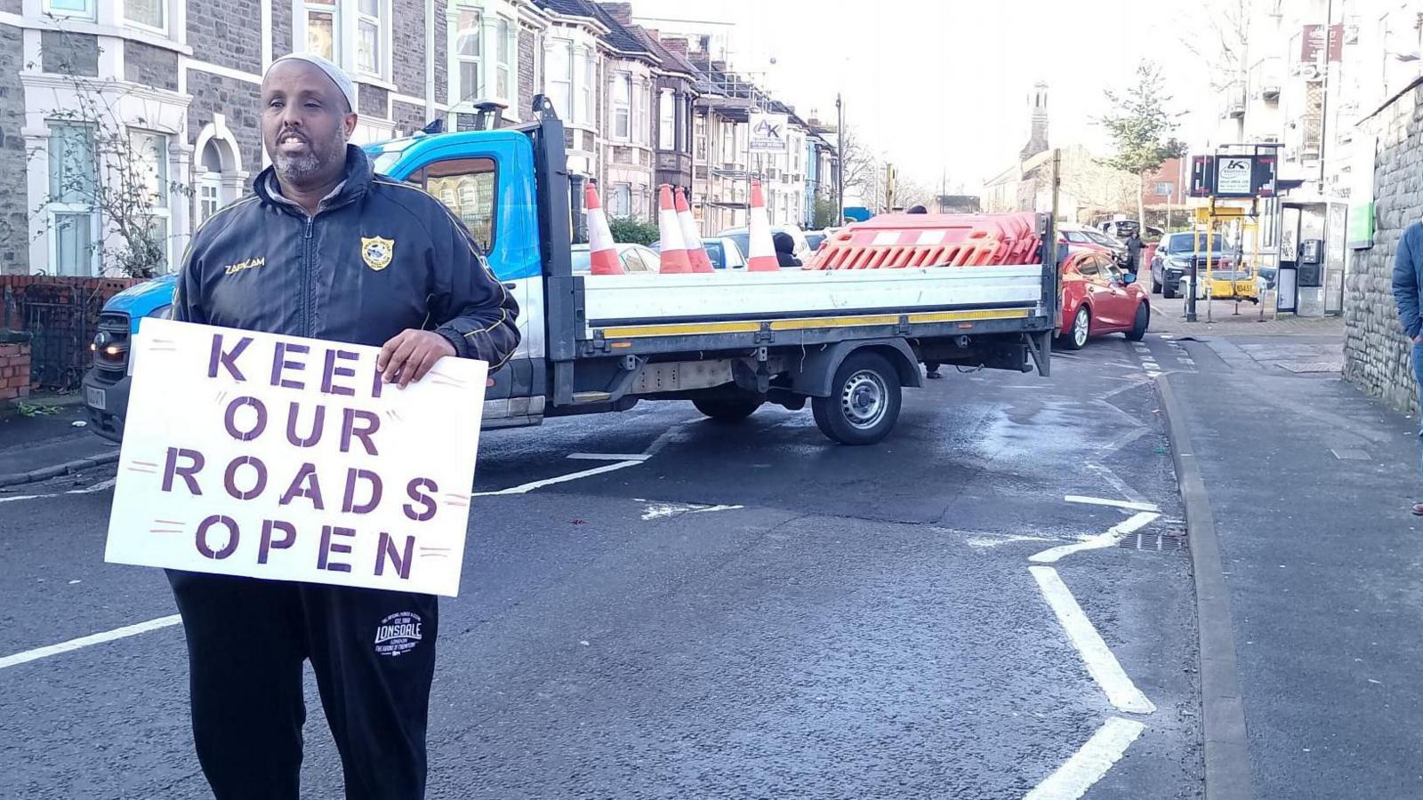 A man standing in the road in front of a truck carrying cones and barriers to stop traffic. The man is holding a sign reading "keep our roads open". 