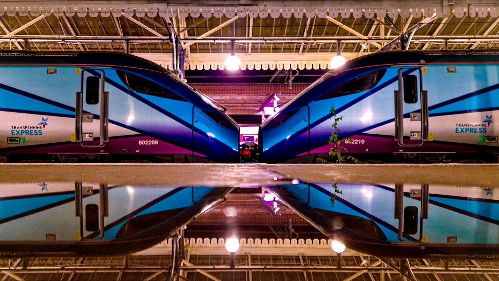 Two train carriages meet on York Railway Station and are reflected in a puddle on the opposite platform