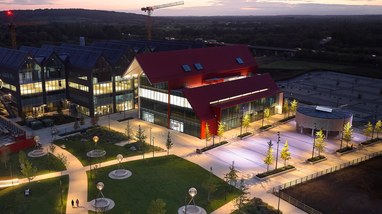 Seen from the air at dusk, the new buildings are lit up from inside. Part of a park can be seen alongside.  There are pale paths cossing the grassed area, with several spherical glass artworks in the park.