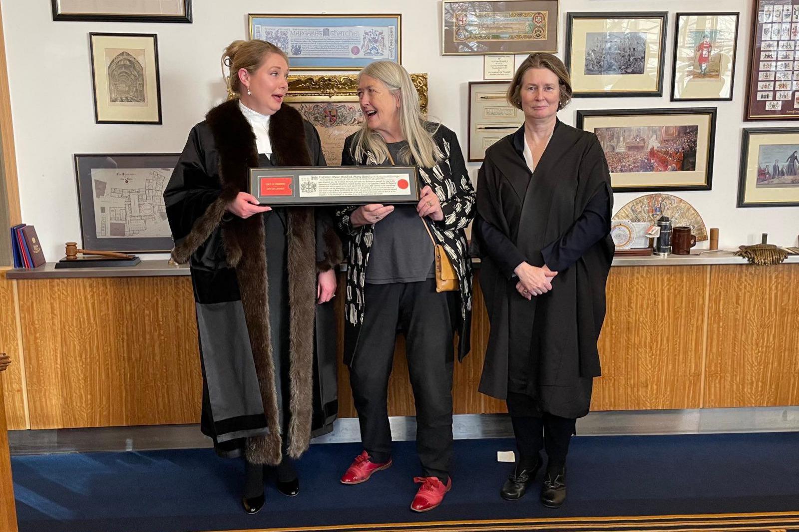Mary Beard stands in between two other women who are wearing black cloaks and gowns. She is being handed a document in a frame by the woman to her left