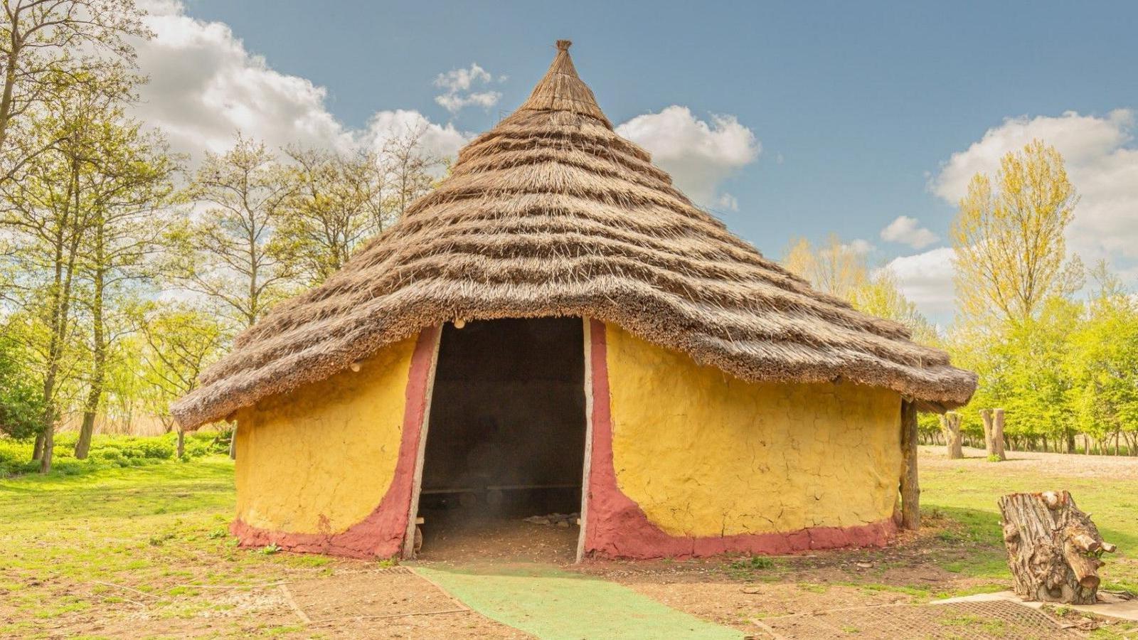 A roundhouse replica with a thatched roof. The walls are made of mud/clay with a wide door-shaped entrance. The exterior wall is painted saffron yellow. There are trees behind the roundhouse.