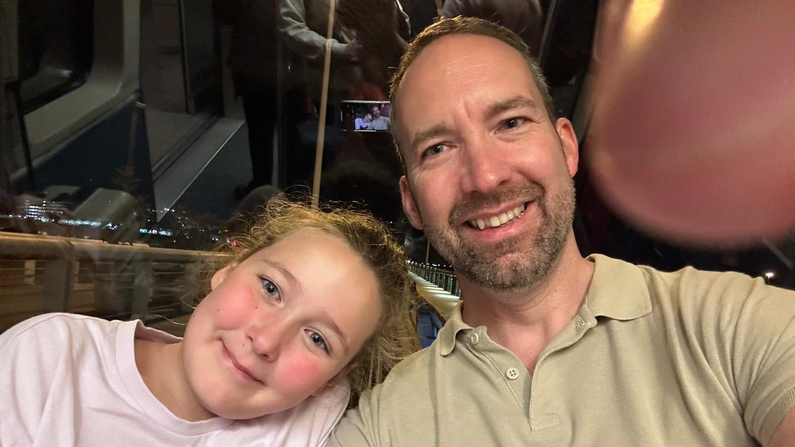 Jonathan with his daughter, both of whom are smiling. They seem to be sat on public transport at night. The picture is a selfie.