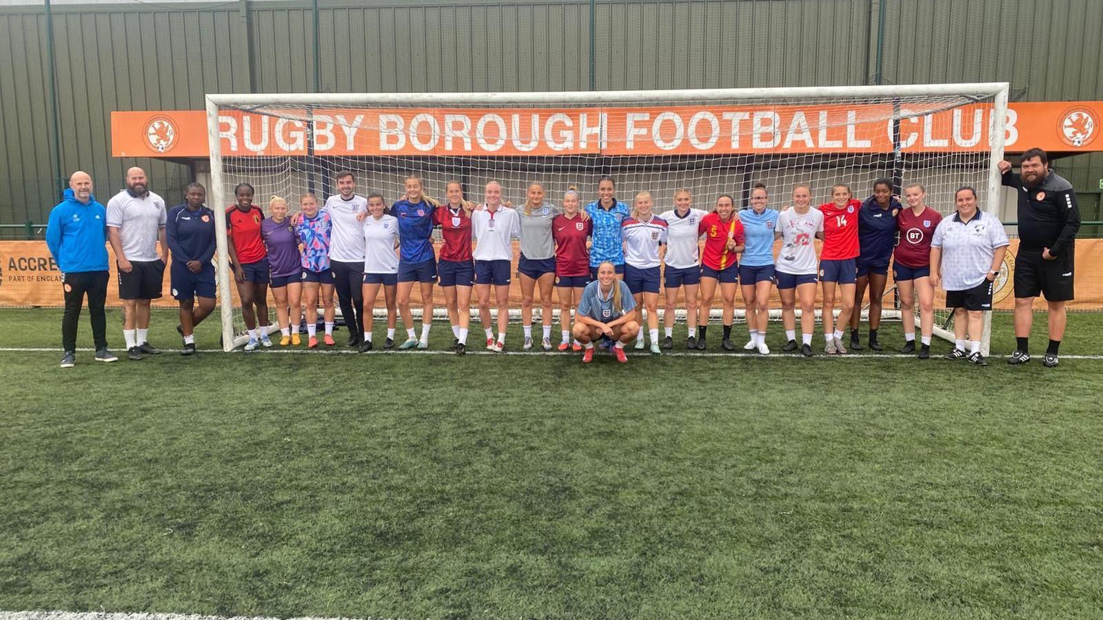 A football team of women wearing England shirts standing in a goal on a football field. Male coaching staff stand either side of the goal posts.