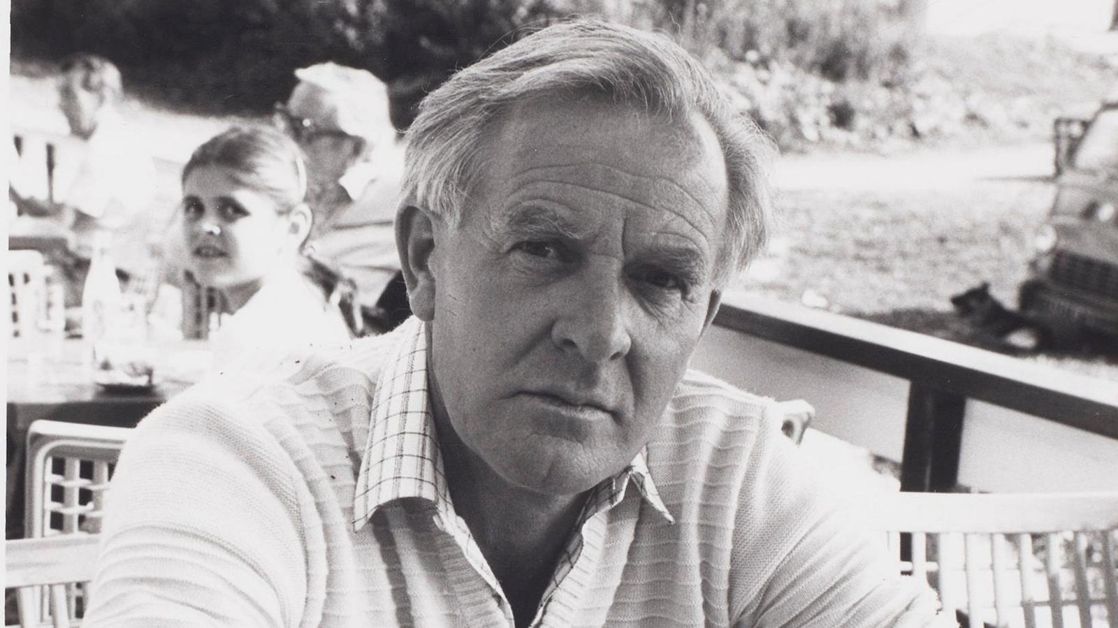 A black and white photo of a man wearing a long-sleeved pale top with the sleeves rolled up. He's sitting at a table and leaning forward on his elbows, and looking, unsmiling, at the camera. There's a wooden railing and some other people sitting at tables behind him.