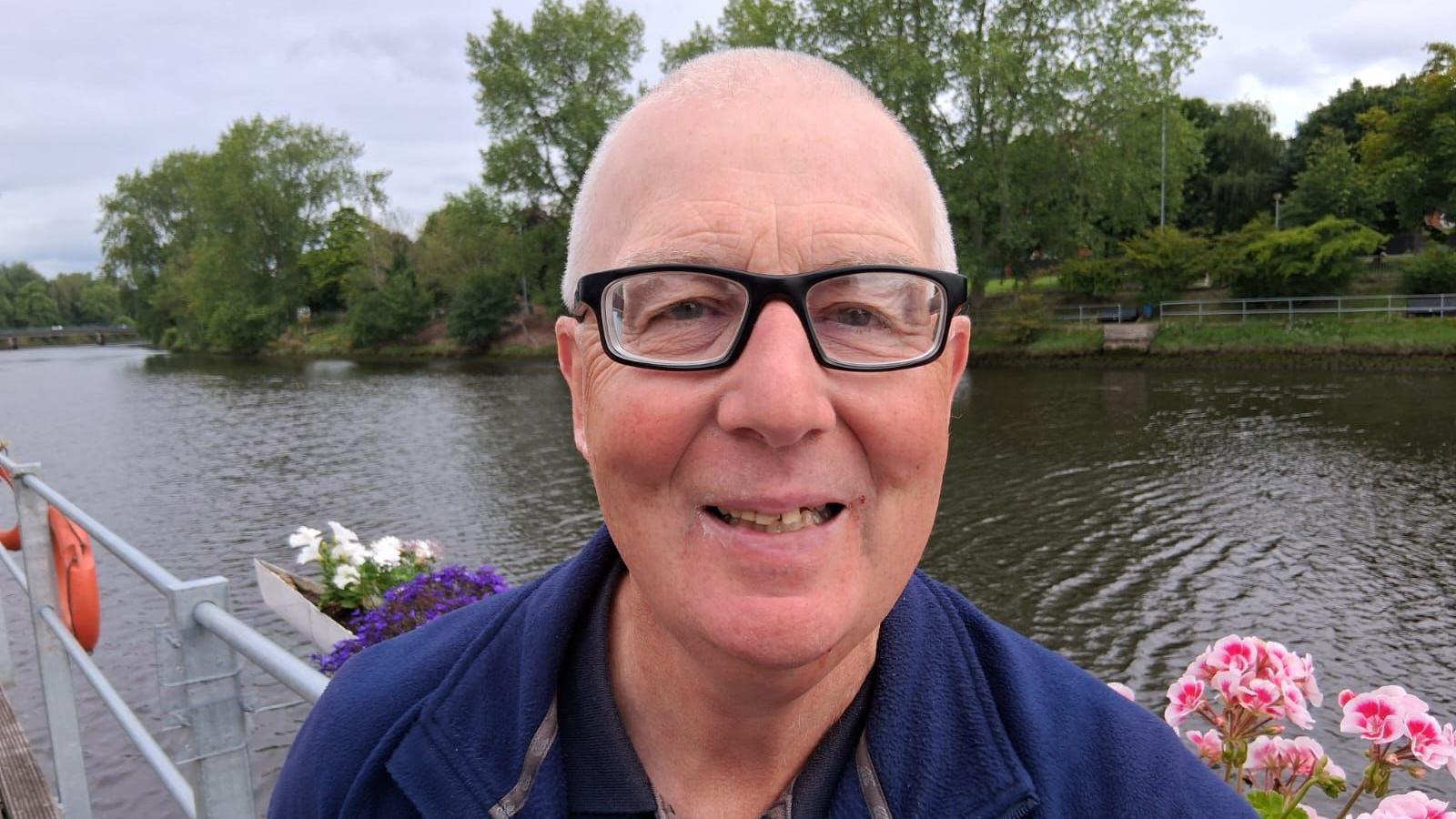 Methodist College Belfast head coach Enda Marron standing in front of the River lagan. There are pink flowers along the grey guard rail. There is a green embankment in the background with green trees.