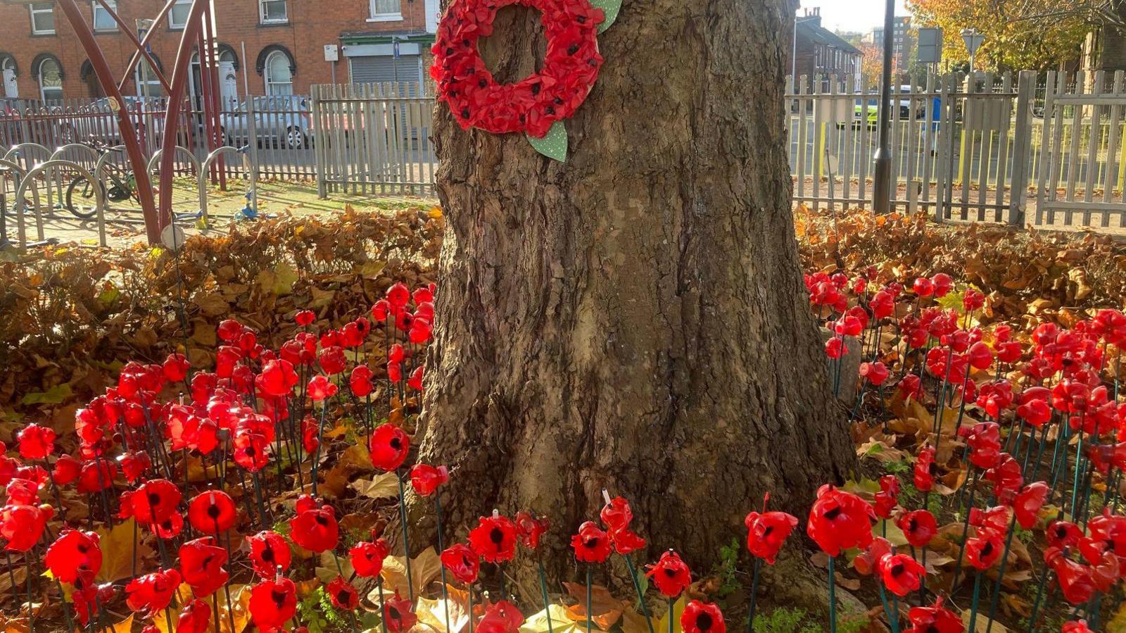 Red plastic poppies on sticks around the base of a tree, which is surrounded by brown autumn leaves. There is a metal grey fence and houses behind and bike racks to the left