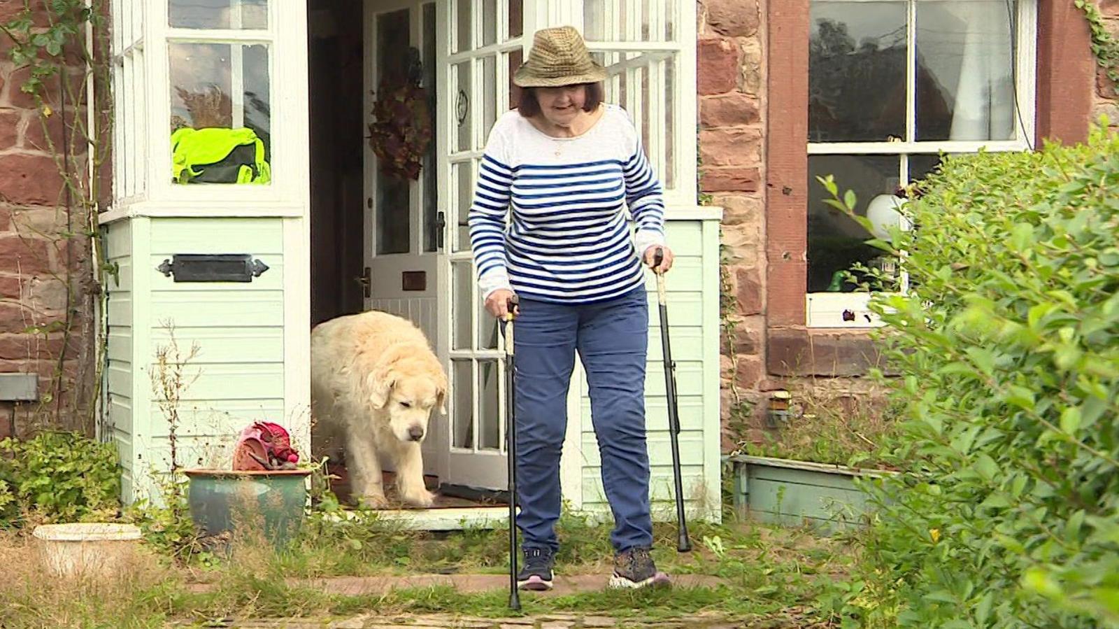 Carol Smithers walking out of her door of her home, into her garden, holding two walking sticks. She is followed by a golden retriever dog.