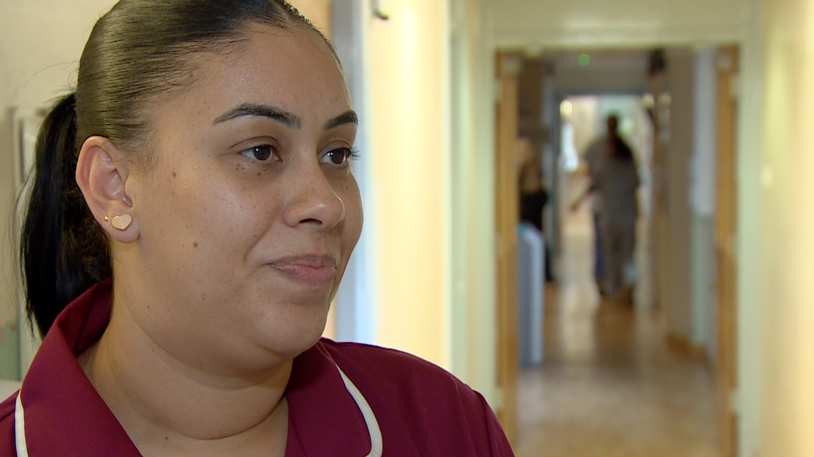 Nadia Tavernier-Gustave, a nurse wearing a maroon tunic in a hospital corridor. She is facing to the right and looks serious