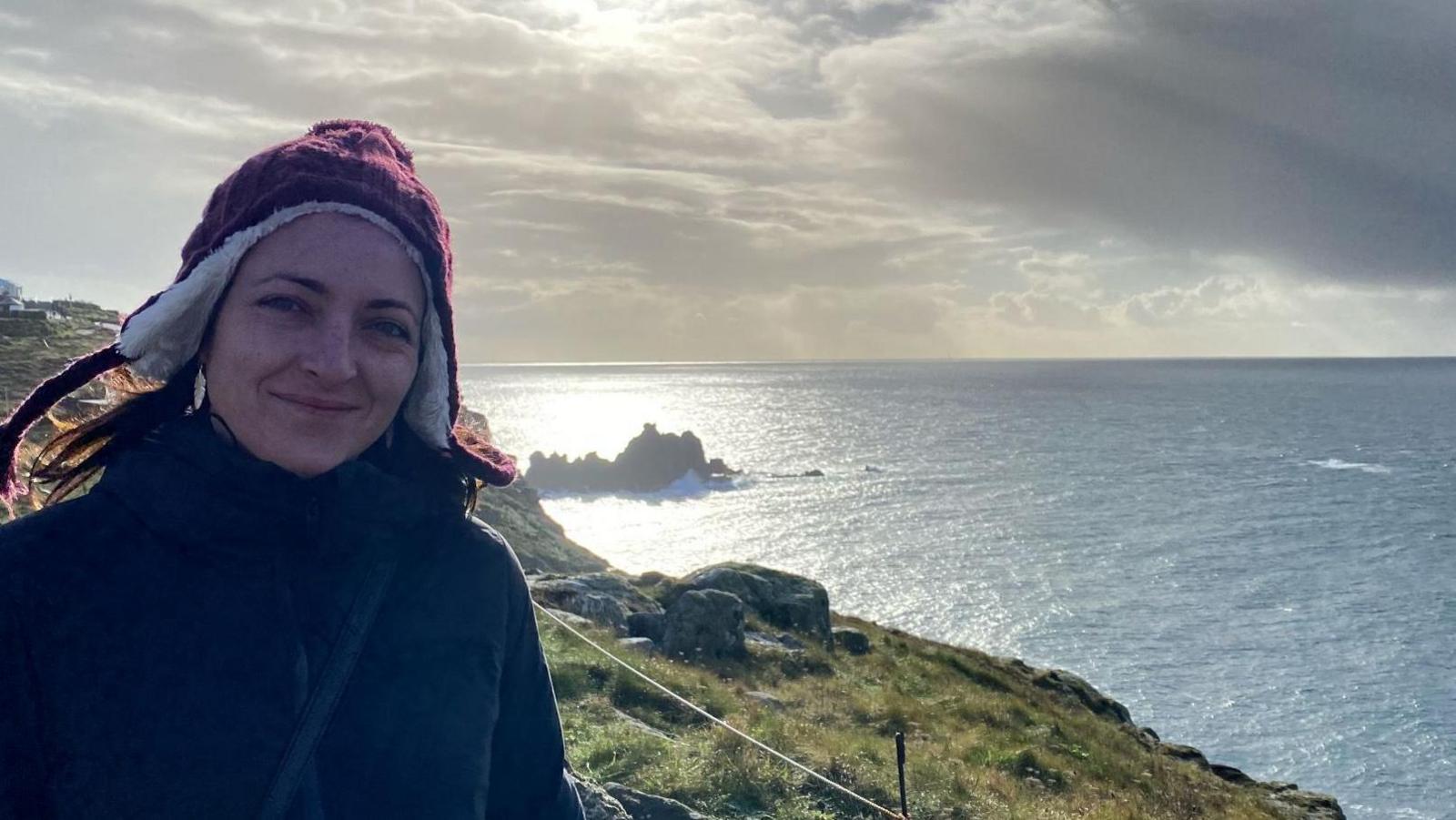 Emily Jones wearing a woollen hat stands on a clifftop path with the ocean behind her