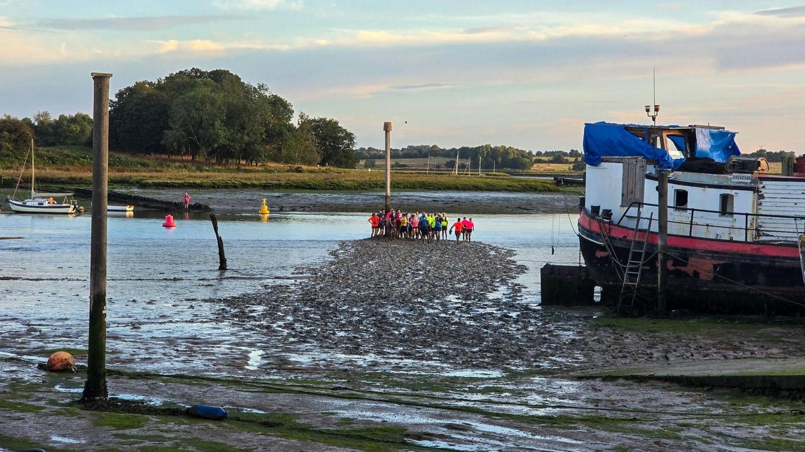 A distant view of the runners gathered together on the river's muddy bank in their brightly-coloured tops. There is an old wooden boat in a state of disrepair to the right.
