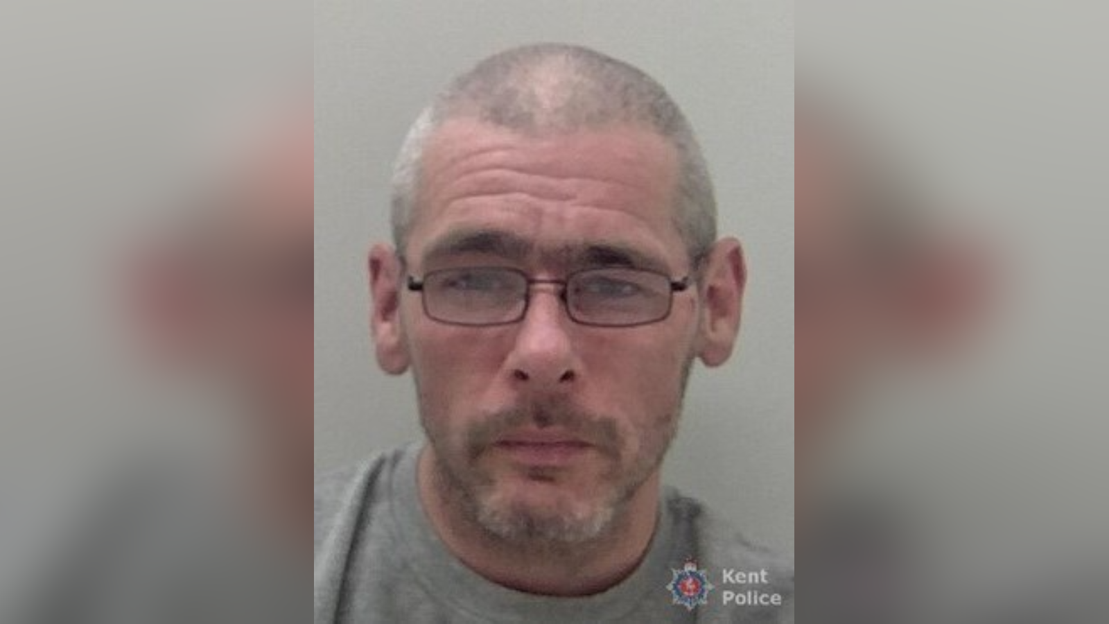 A police mug shot of a man with a shaved head and glasses.