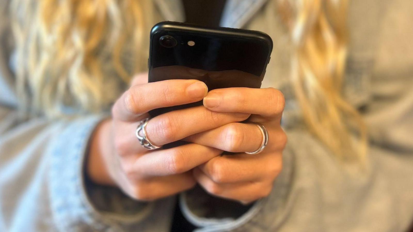 A close up of hands holding a black mobile phone. The person is wearing two silver rings.