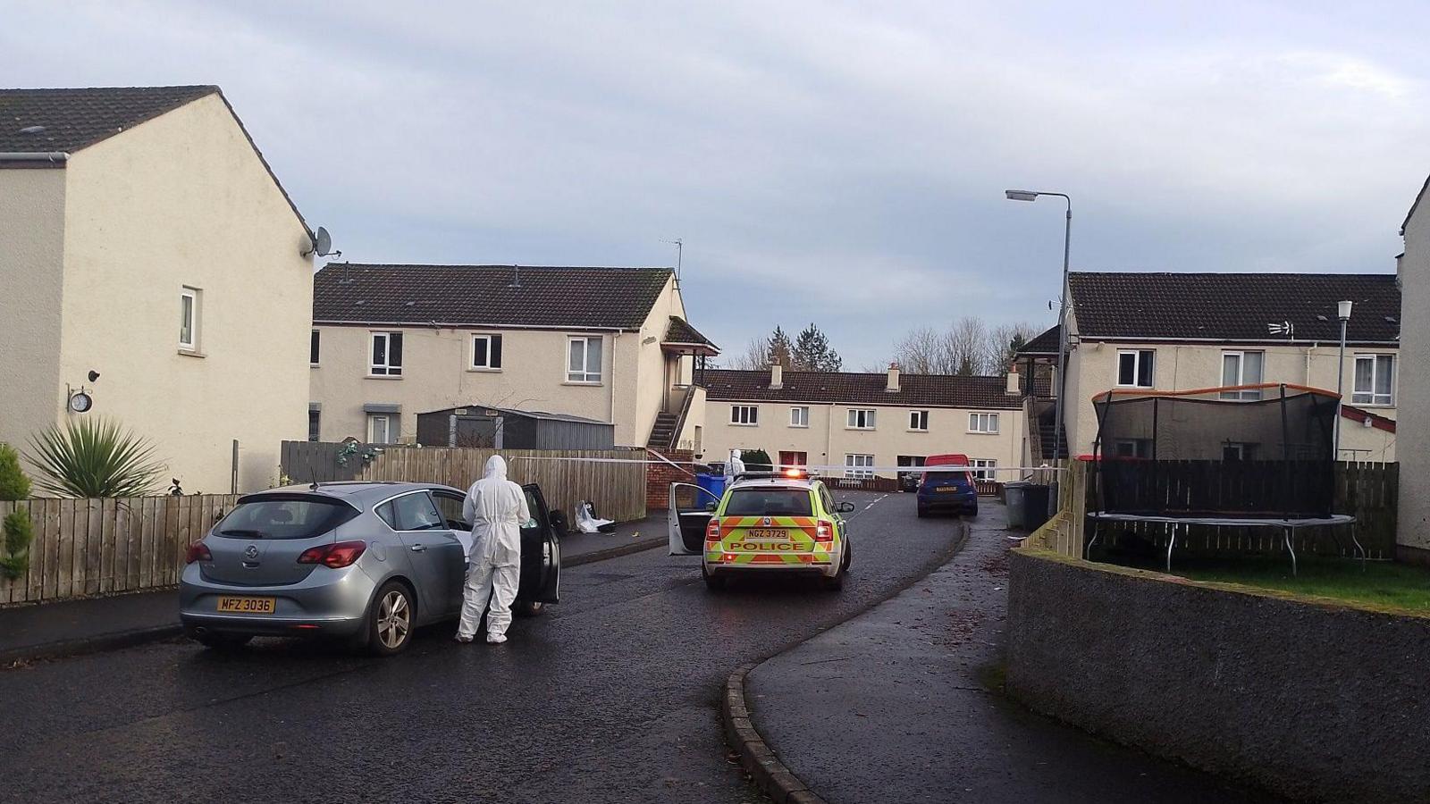 Officer in a forensic suit gets out of a silver unmarked car with a police are in front of it and a cordon across the street in a housing estate. One of the houses has a trampoline in the back garden