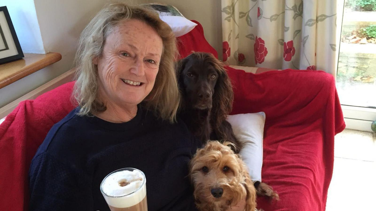 Lynne Adams smiling into the camera holding a coffee on the sofa, next to her two dogs