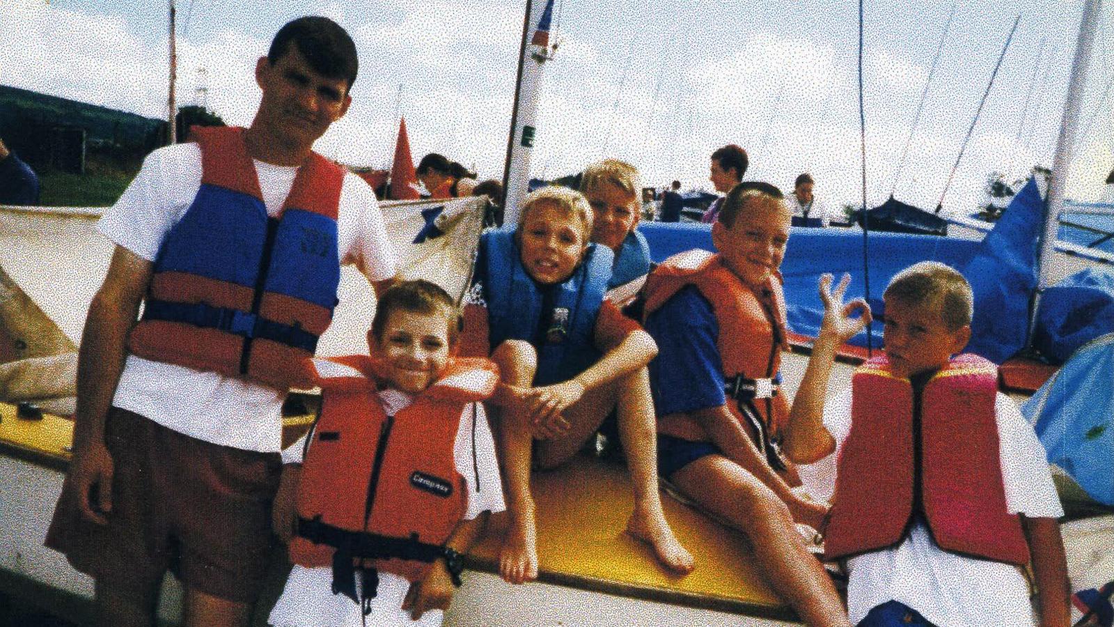 Anton sits on a boat with the Amoss family. They are all wearing life jackets  and there are people stood behind them with the masts sticking in the air from other boats.