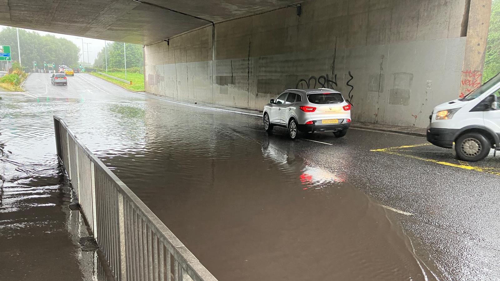 A car and a van try to drive through a flooded underpass with a concrete wall in the background and a metal fence in the foreground