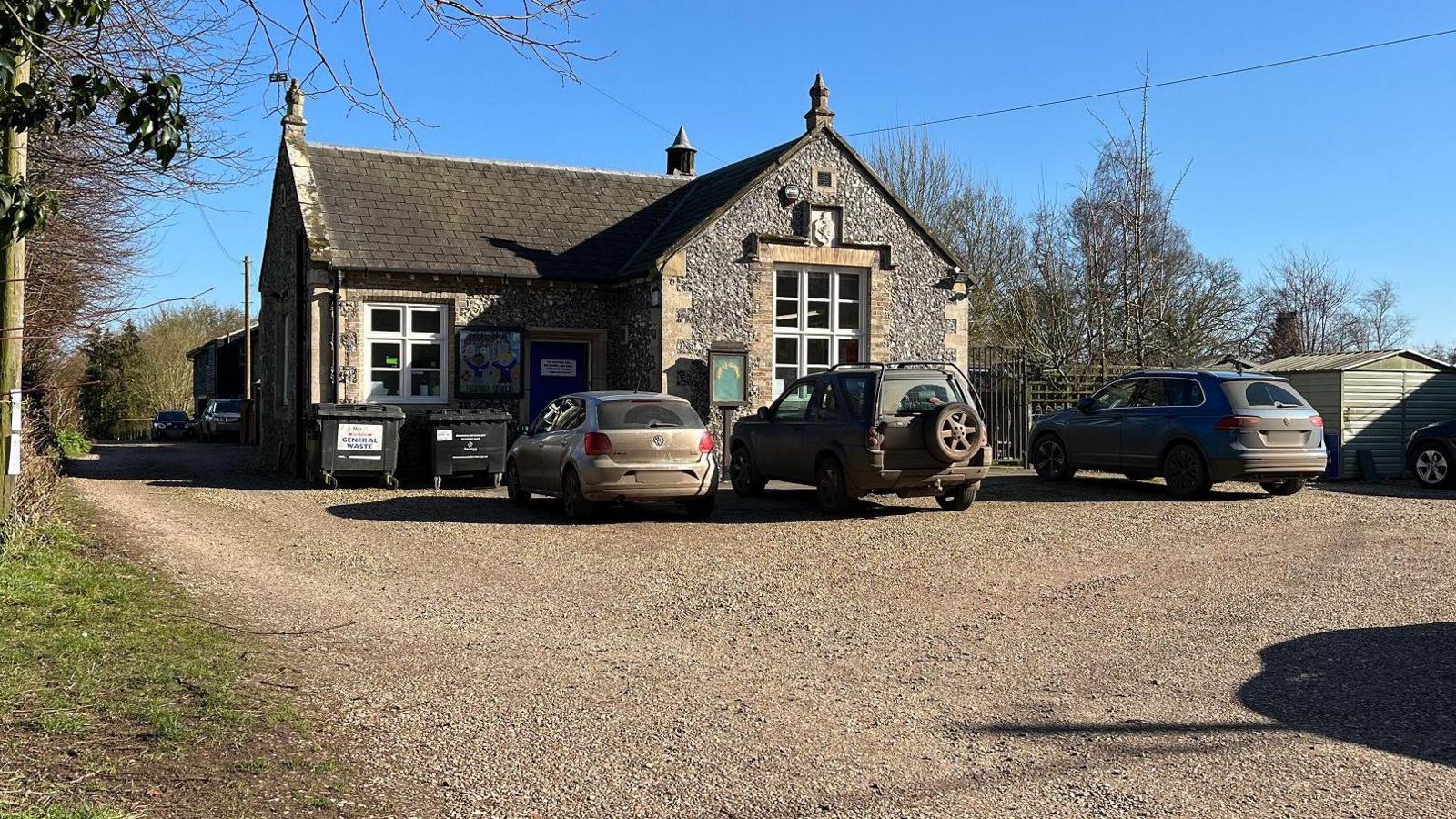 Great Dunham Primary School is a small flint-covered building. Cars are parked in front of it and a gravel drive, to the left of the picture, runs past it. 