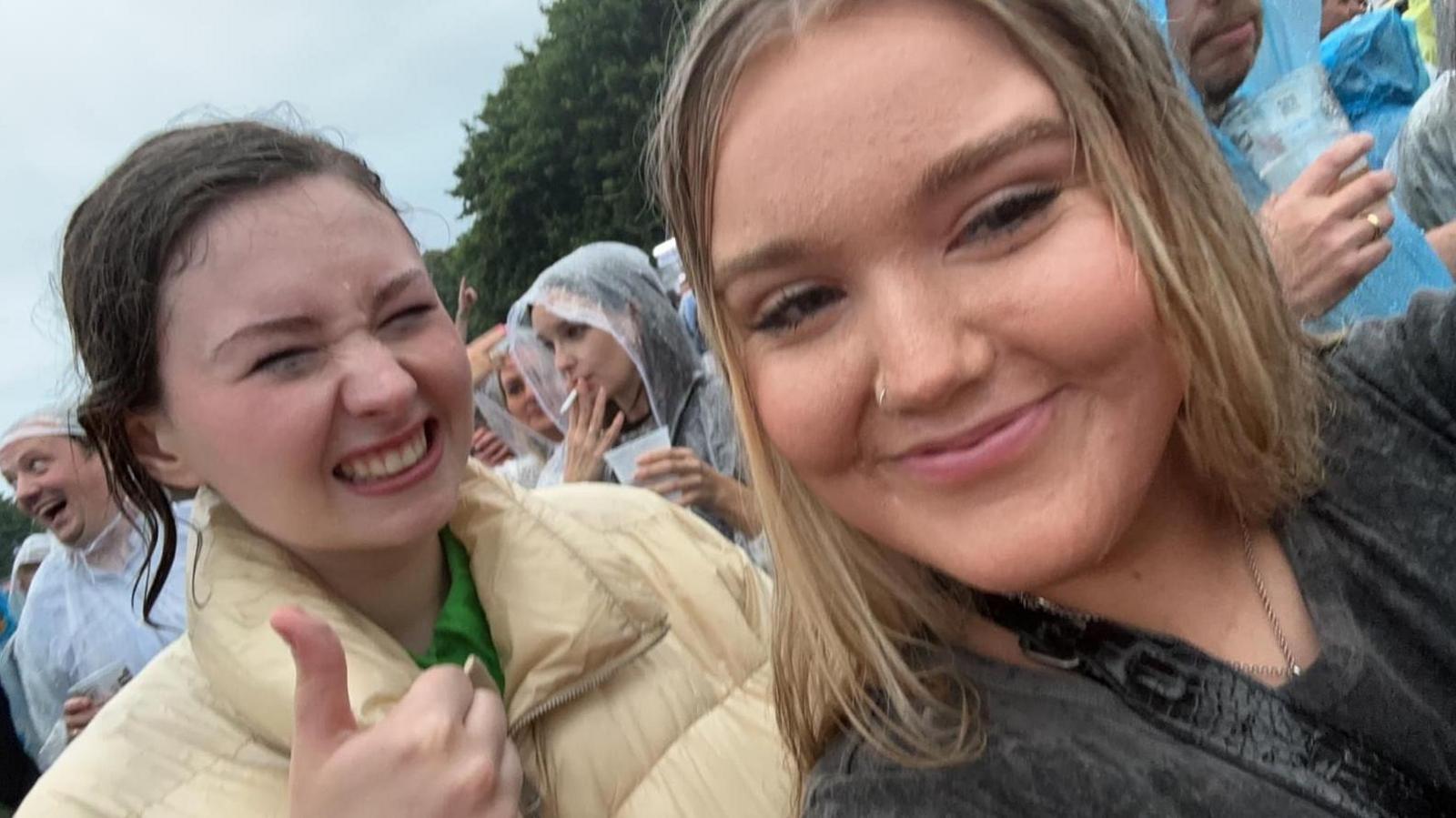 Grace is at the front of the picture, taking the selfie. She has blonde hair and is smiling at the camera. She has a nose ring and is wearing a black top. Her friend is in a yellow puffer jacket and is giving a thumbs up while grimacing in the rain. There are a number of people behind them wearing ponchos.