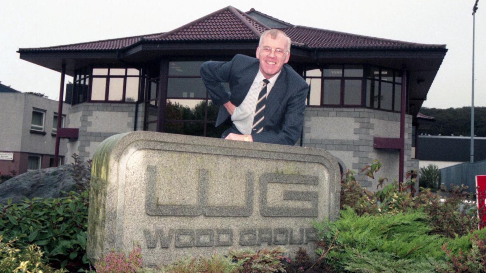 Sir Ian Wood in front of the Wood Group headquarters. He is lying across a concrete sign, smiling.