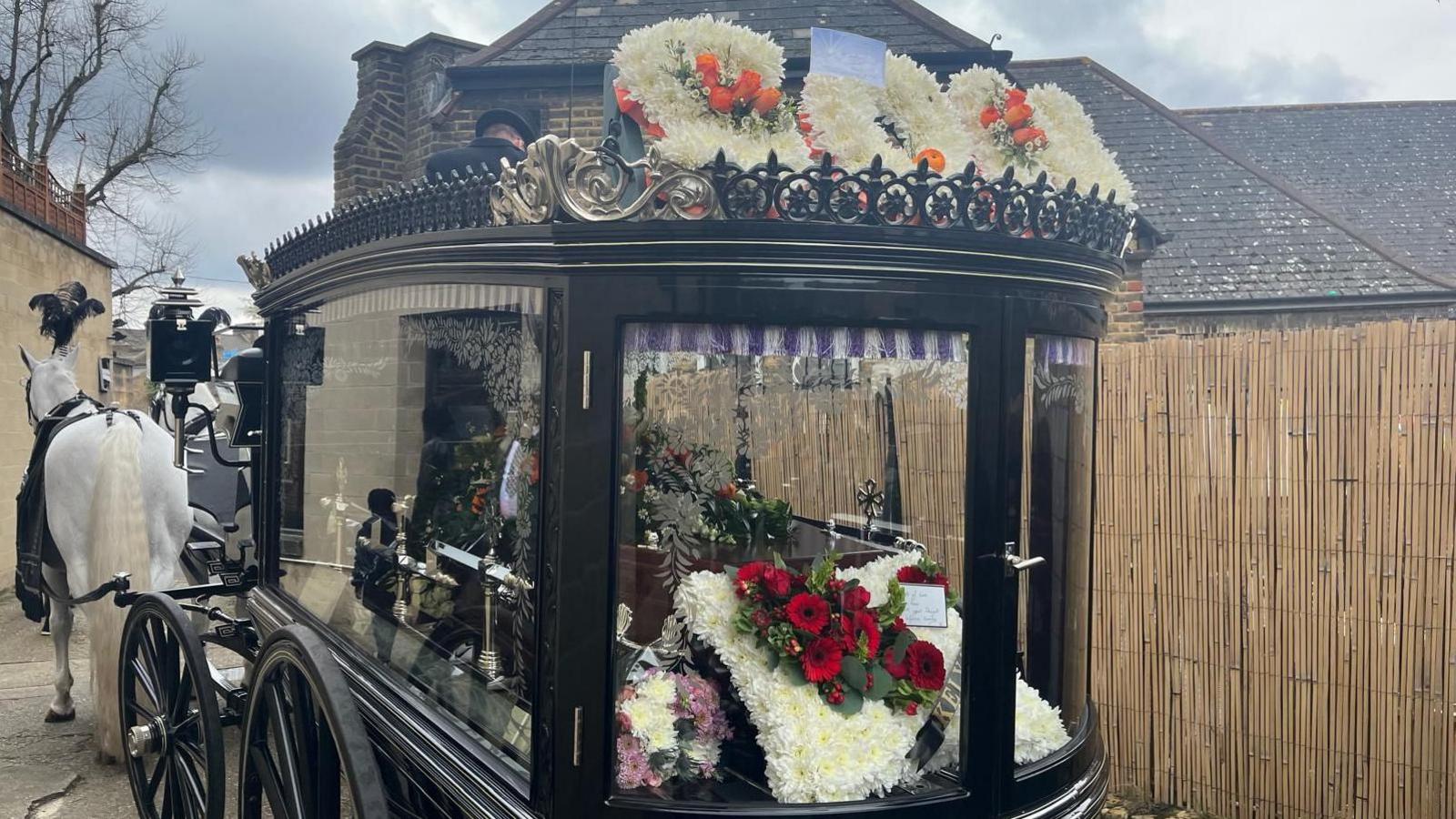 The coffin, surrounded by white and red flowers hearse, being pulled in a carriage by white horses. On top of the carriage is the word 'son' in white flowers with orange flowers dotted around. 