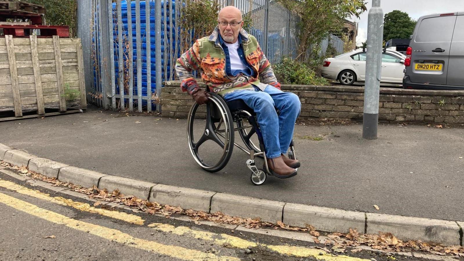 Picture shows David Burdus who is sitting in a wheelchair. He is on a pavement with the kerb showing.