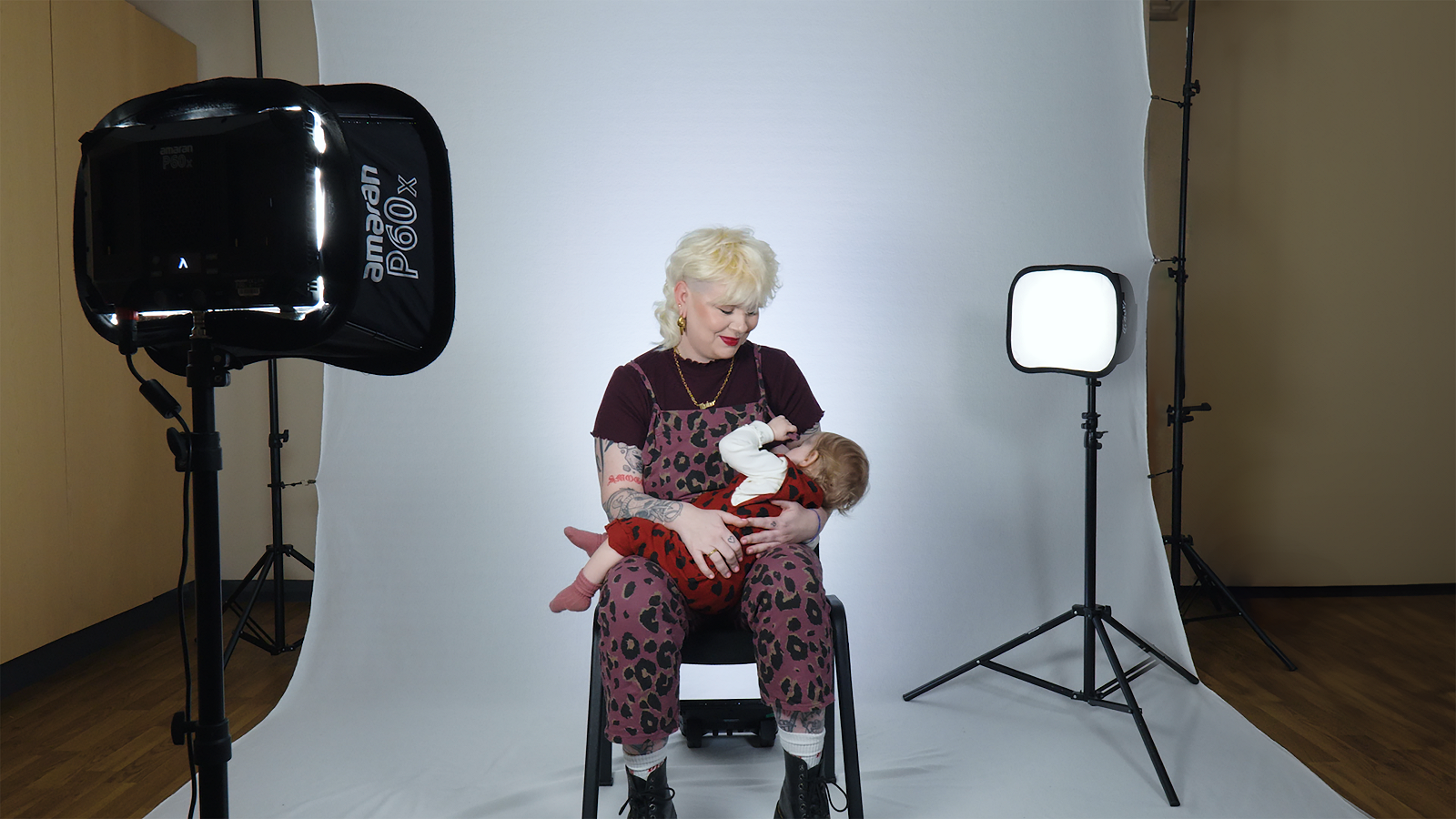 Emma Cooksey with her baby at the campaign photoshoot. Emma is seated and has blonde hair. She is wearing a maroon top and maroon dungarees. She has tattoos on her arms. Her child has fair hair and is wearing a red outfit. She is lying in her mum's lap. They are sitting in front of a white background with a camera on the left-hand side of the photo. A light box on top of a tripod can be seen on the right.