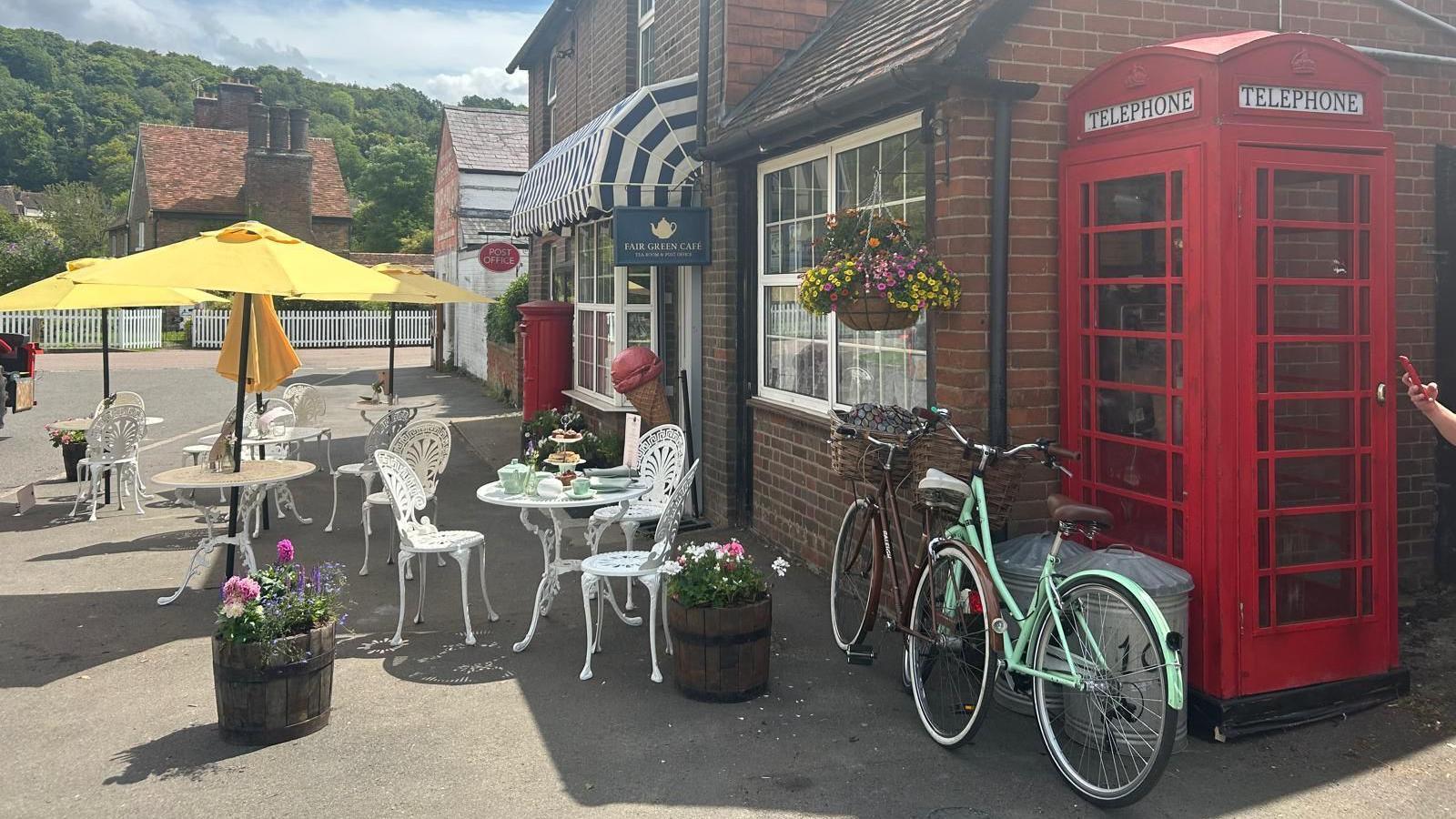 Outdoor seating area of a café with white iron tables and chairs, a large yellow umbrella, a red British telephone box, bicycles, and potted flowers.