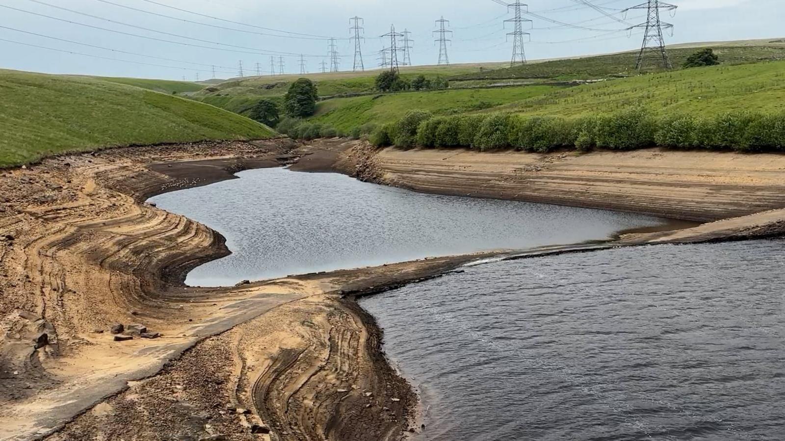 Low water levels visible at a reservoir, with muddy banks to either side of water. Above is grass and trees, with electricity pylons on the horizon and a blue sky above.