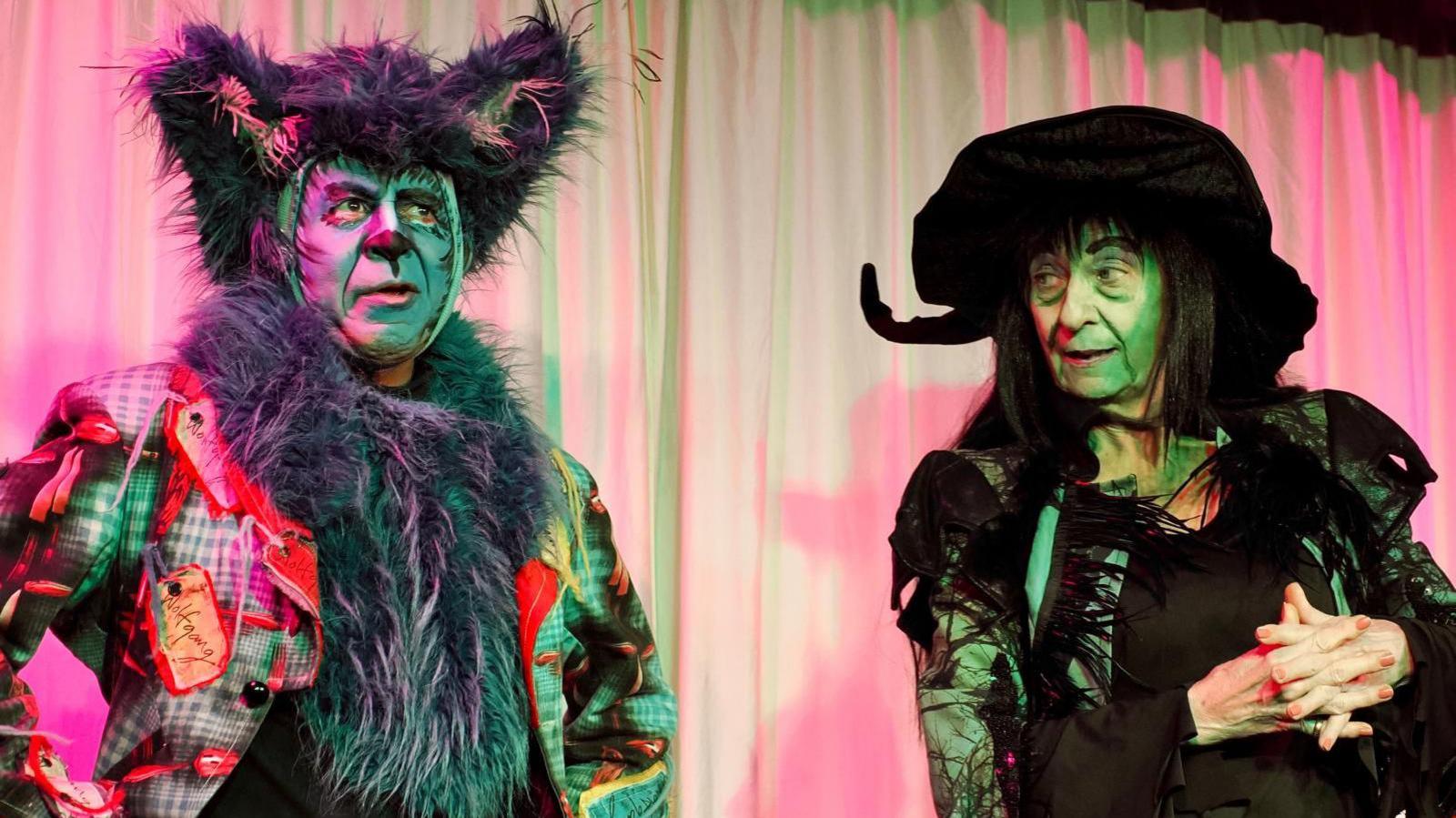 A man and woman stand on stage beneath fluorescent lights, dressed as a wolf and the Wicked Witch of the West from the Wizard of Oz.