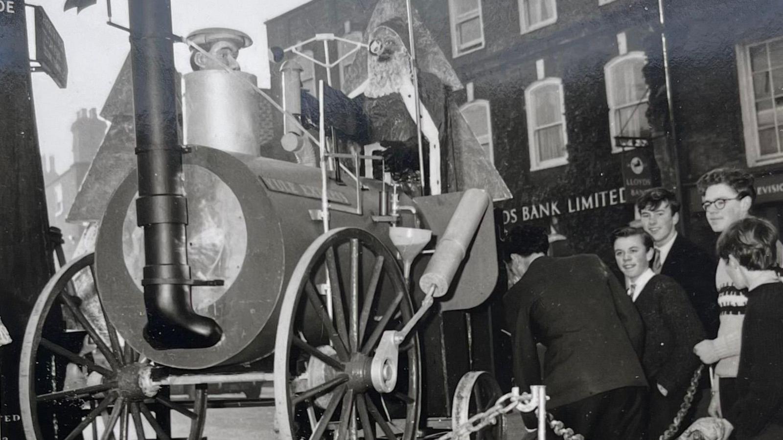 A black and white photo of four teenage boys - mostly smiling - and standing next to a chain which has fenced off a replica of the engine, with a model of Father Christmas at the controls. Lloyds bank sign is in the background.