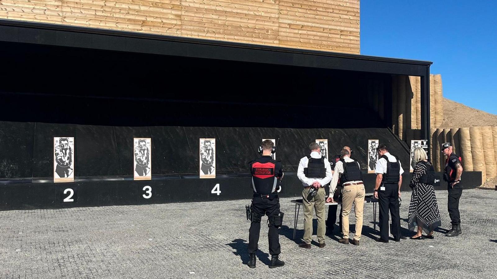 Five men and a woman all wearing bulletproof vests have their backs to the camera as they look at seven targets with men on them. Three of the targets are labelled 2, 3 and 4.