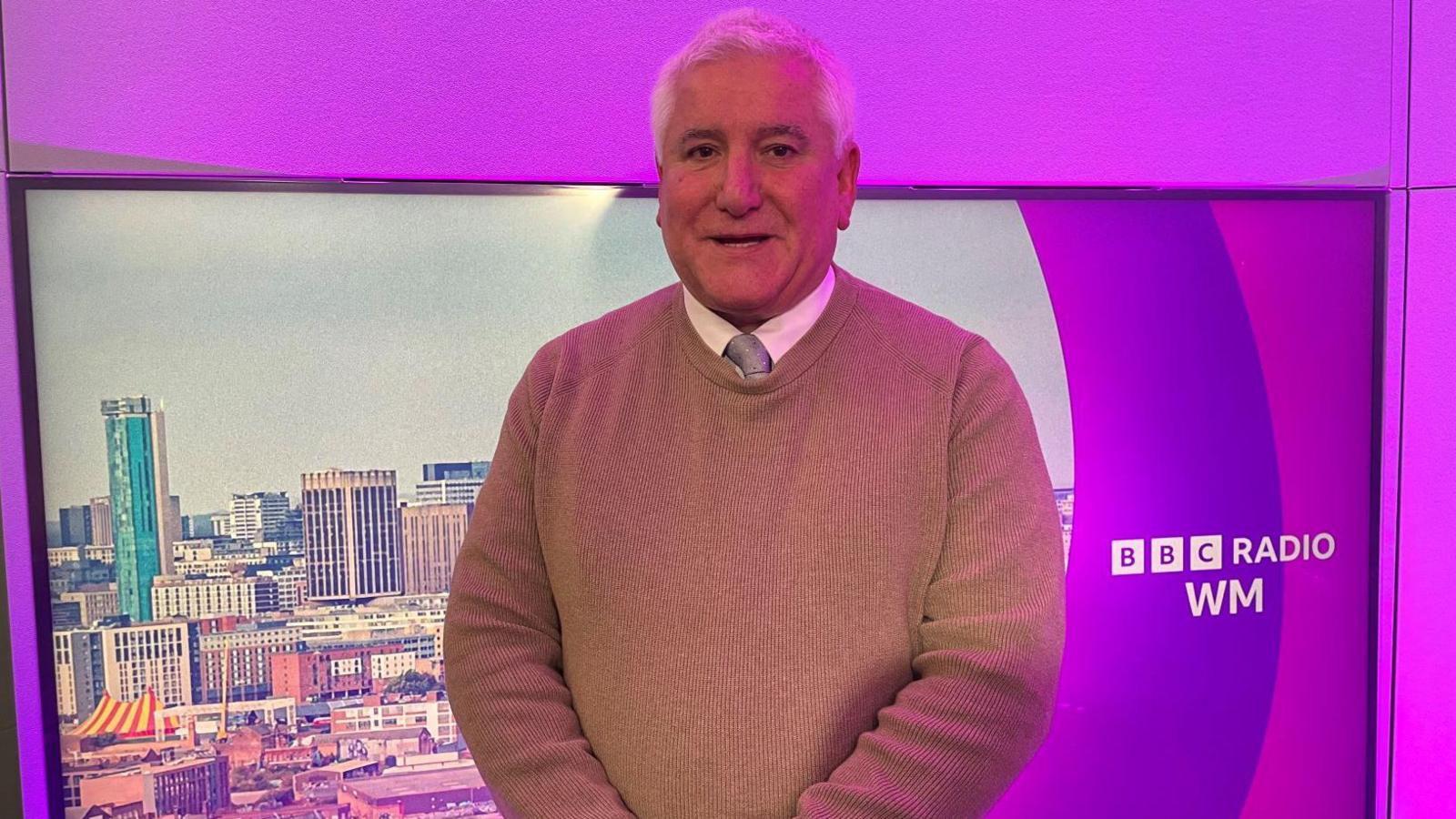 A man stands in front of a pink screen. He has grey hair and is wearing a brown jumper, a white shirt and a silver tie. The pink screen says "BBC Radio WM" on it. 