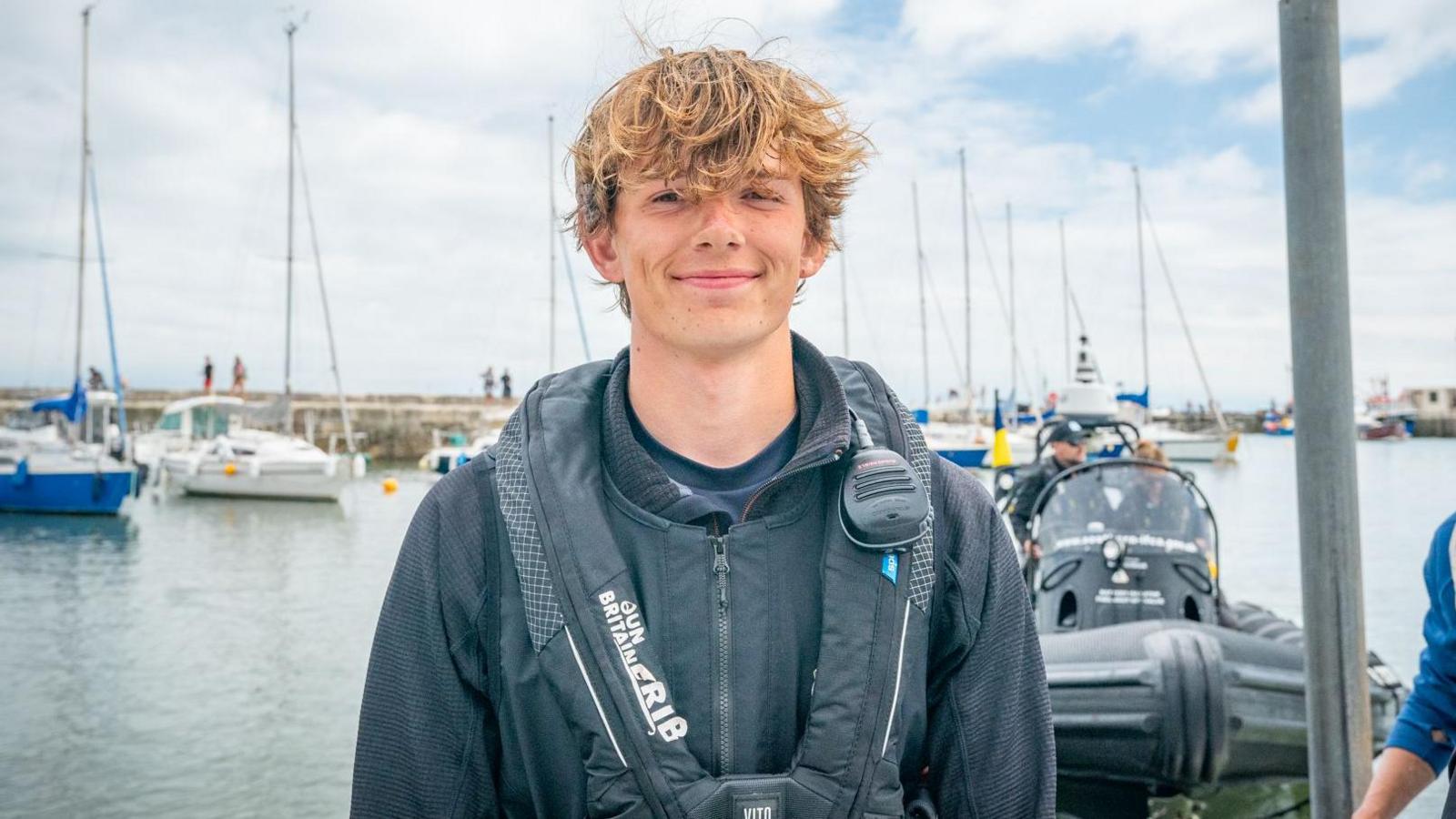 Harry Besley wearing a black waterproof jacket and a life jacket, with a radio strapped to his shoulder. He has short brown windswept hair and is standing on a dock in Lyme Regis harbour, with sailboats visible in the background