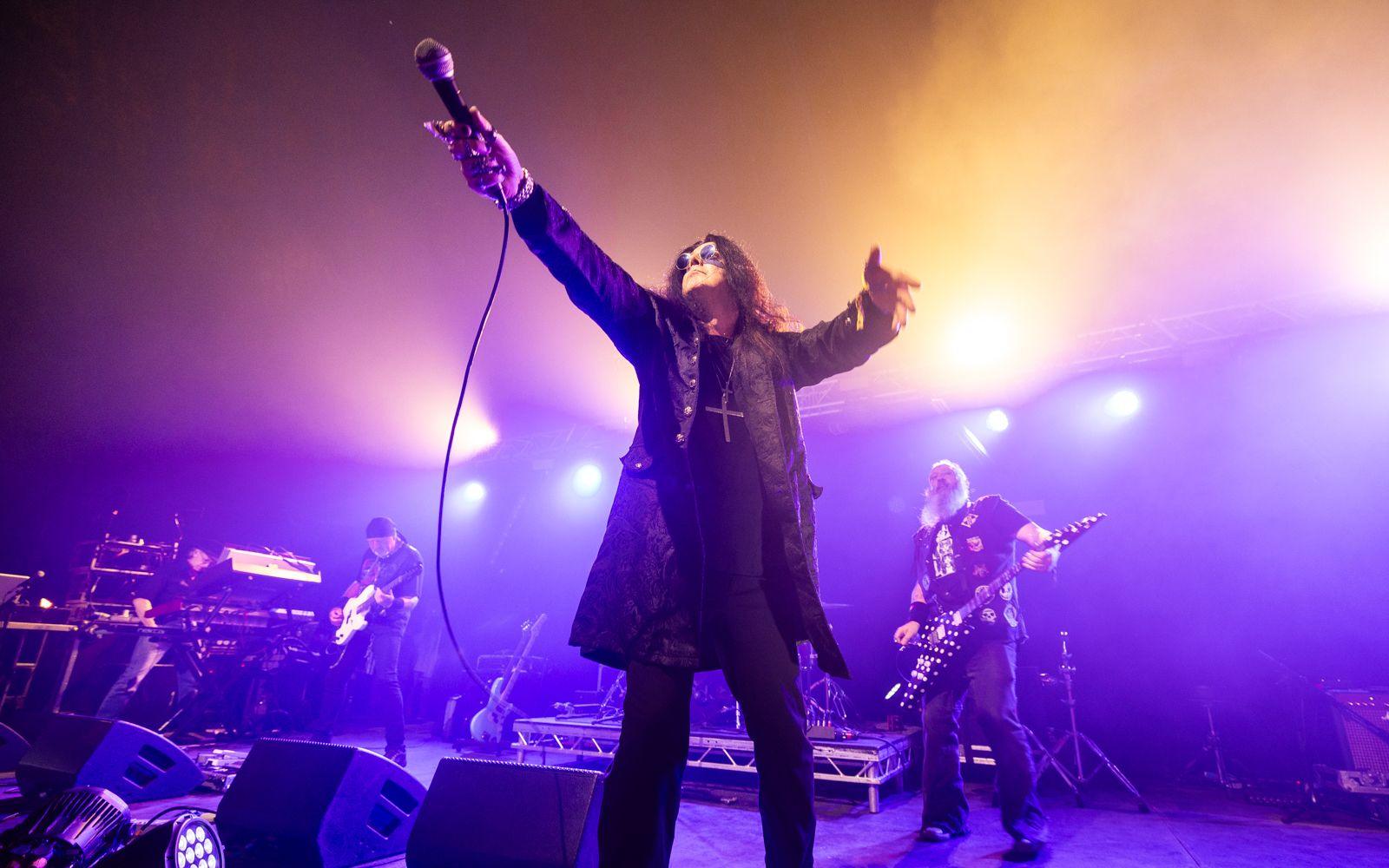 Singer dressed in the style of Ozzy Osbourne - dark jacket, round sunglasses and crufix necklace - holds a microphone towards the audience from a purple lit stage