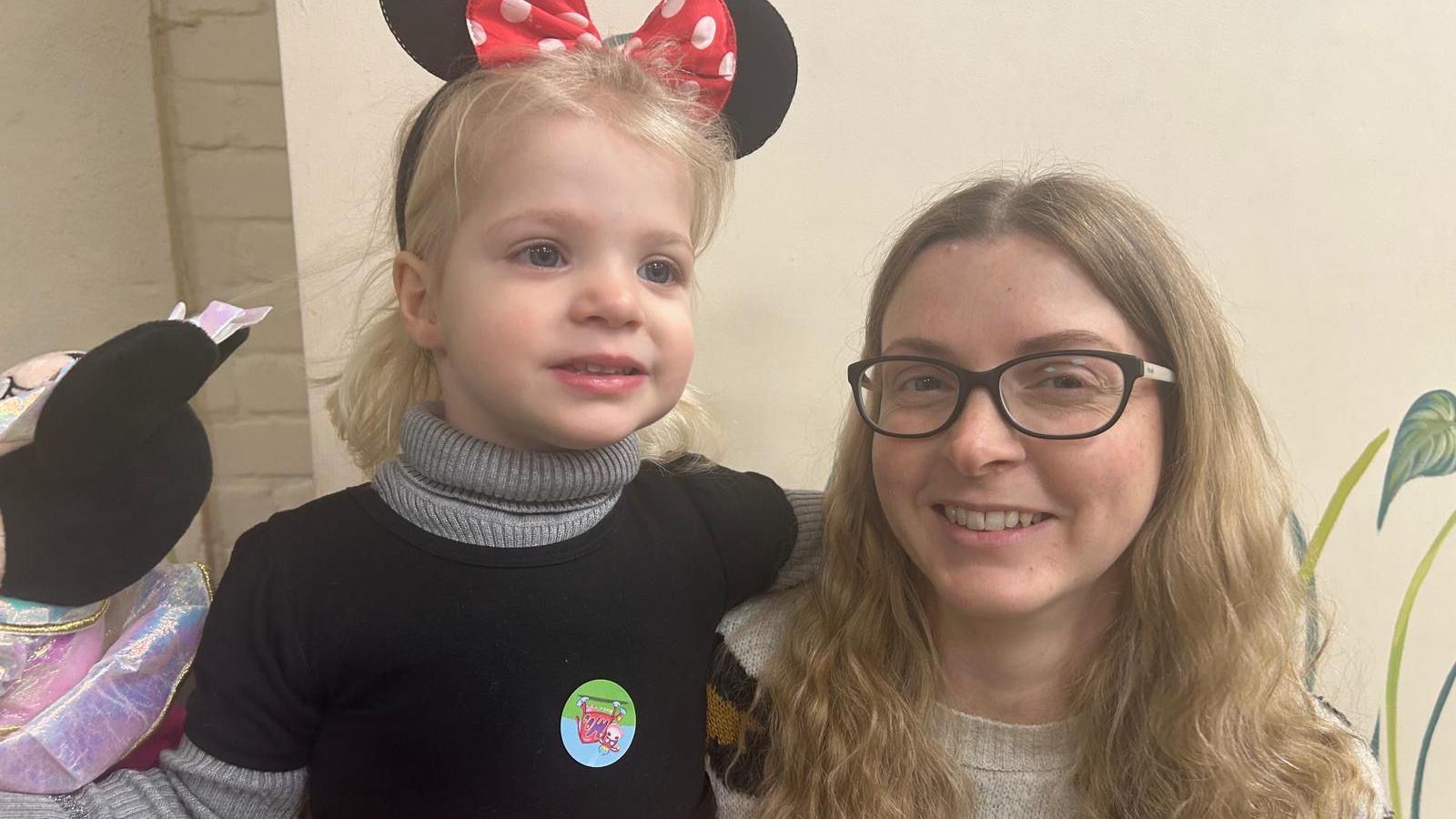 Rachel May with her daughter. Her daughter has black Mini Mouse ears on and is wearing a black top. Rachel is wearing glasses with a grey jumper.