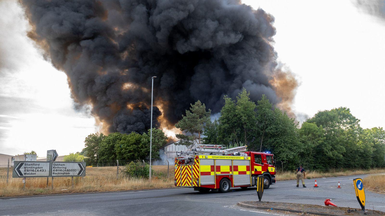 A fire engine parked on the road and there is a large plume or grey/black smoke behind it 